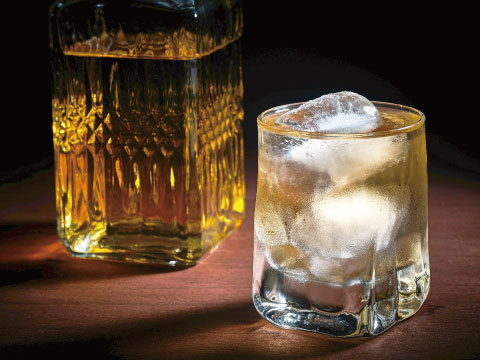 Glass of whisky on the rocks beside a cut-crystal decanter of amber whisky on a wooden table against a dark background