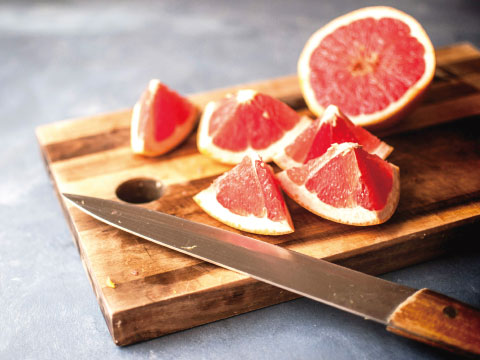 Fresh pink grapefruit cut into wedges and halves on a wooden cutting board with a kitchen knife on a gray table