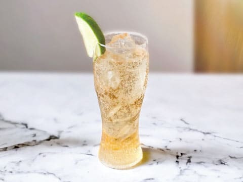 Sparkling golden ginger ale over ice in a tall glass garnished with a lime wedge on a white marble table