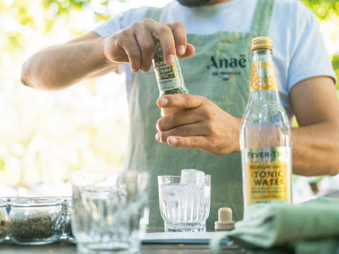 A bartender in a green apron pouring Fever-Tree tonic water into a glass of ice outdoors, with another tonic bottle on the table