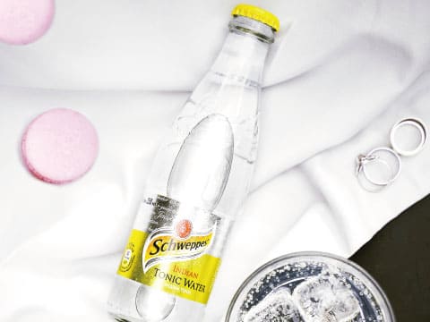Bottle of Schweppes Indian Tonic Water on white linen beside a glass of ice, macarons and silver rings