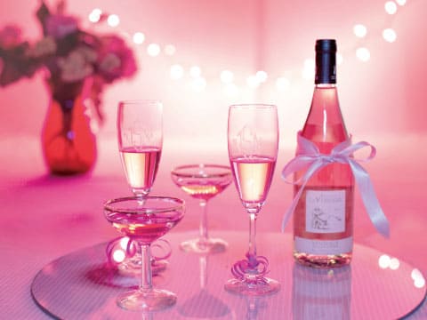 Bottle of rosé wine tied with a white ribbon beside pink-tinted coupe and flute glasses on a glass table with soft pink lights