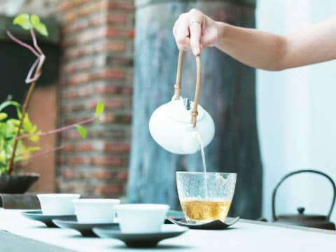 A hand pouring golden tea from a white ceramic teapot with a bamboo handle into a clear glass, with small teacups on a table beside a brick wall