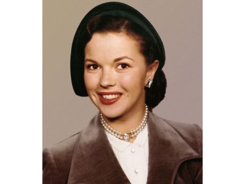 Color portrait of Shirley Temple Black, the American actress who later became a U.S. diplomat, wearing a dark green hat, pearl necklace and brown coat