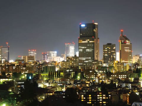 A Japanese city skyline at night, with illuminated high-rise towers rising above the warm lights of a dense residential area under a dark sky