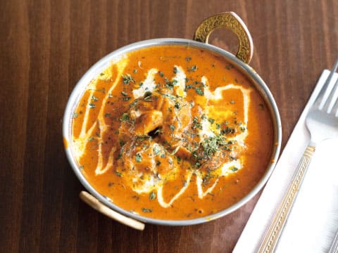 Creamy butter chicken curry with a swirl of cream and dried herbs in a small serving bowl with brass handles, a mild spiced pairing for the East India cocktail