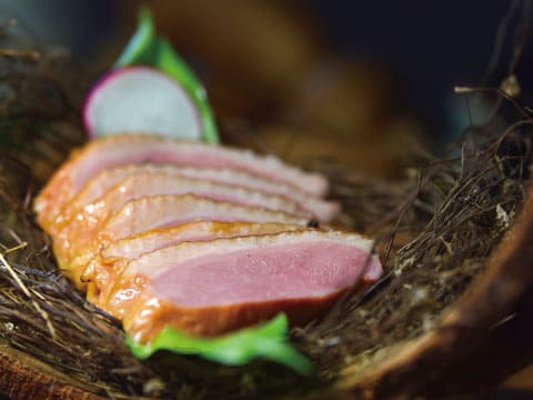 Slices of pink smoked duck breast with a golden glazed edge, arranged on a bed of dried hay with a radish slice and fresh greens