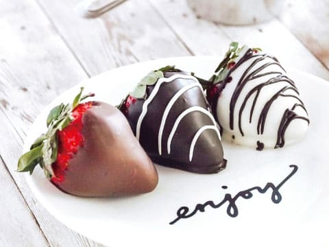 Three chocolate-dipped strawberries in milk, dark and white chocolate with drizzled stripes on a white plate with the word "enjoy"