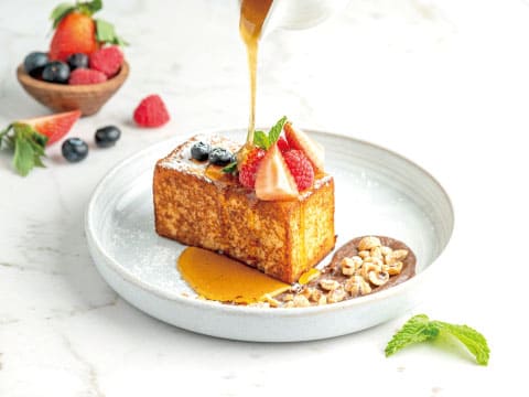 Thick-cut French toast topped with strawberries, raspberries and blueberries, with syrup being poured over it and crushed nuts on a white plate