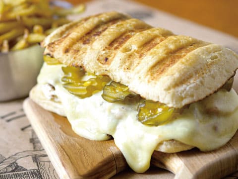 Pressed Cuban sandwich with gooey melted cheese and pickles on a wooden board, served with a side of French fries