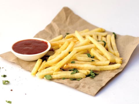 Golden French fries sprinkled with fresh herbs on brown parchment paper, served with a small bowl of ketchup on a white background