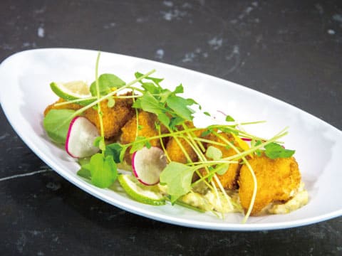 Golden breaded white fish bites topped with microgreens, radish slices and lime on a creamy sauce in a white oval dish