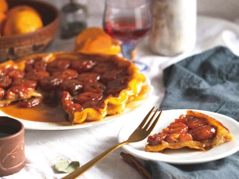 A glossy upside-down tarte tatin topped with caramelized fruit halves, with one slice served on a small plate beside a gold fork and a glass of rosé wine, a dessert pairing for the Apple Pie cocktail