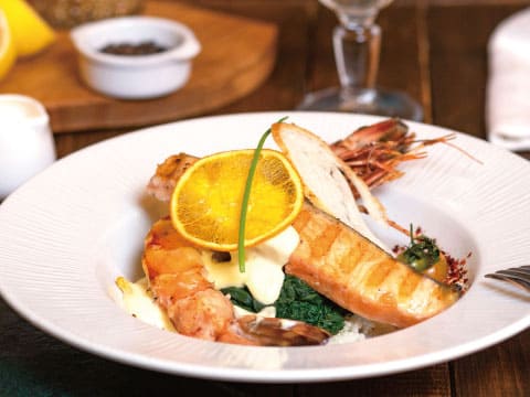 Grilled salmon fillet and prawns served on spinach with a creamy sauce, garnished with an orange slice in a white bowl on a wooden table