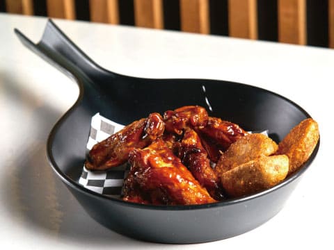 Sticky glazed chicken wings with seasoned potato wedges served in a black skillet-shaped dish lined with checkered paper