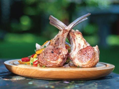 Two grilled lamb chops with crossed bones, seared golden brown and served with sautéed peppers on a wooden plate outdoors, a classic mint-friendly pairing for the Green Hat cocktail