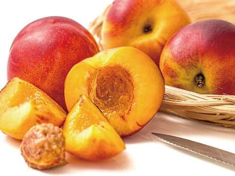 Fresh yellow peaches, whole and sliced, with a peach pit and a knife beside a wicker basket on a white background