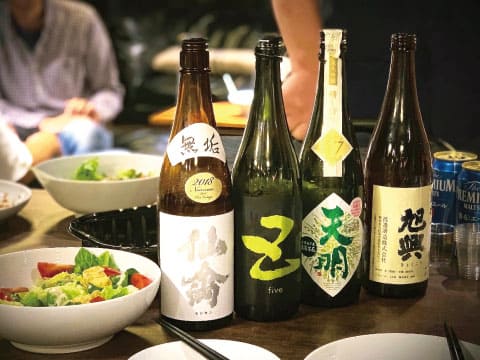A row of Japanese sake bottles with bold calligraphy labels lined up on a dinner table with salad bowls and chopsticks during a casual gathering
