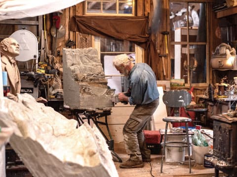 Gray-haired sculptor carving a large block of stone in a cluttered artist’s workshop filled with tools, sculptures and a wood stove