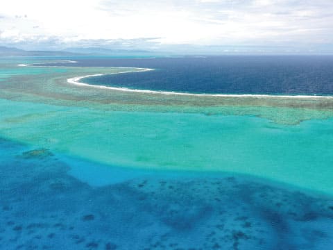 Aerial view of a coral reef with turquoise lagoon waters, a white line of breaking waves and the deep blue ocean beyond