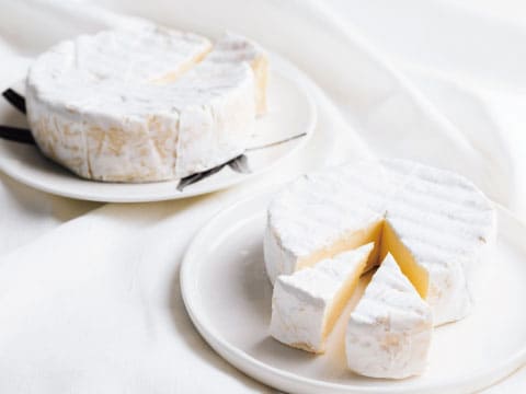Two wheels of soft white-rind brie cheese on white plates, one cut into wedges to reveal its creamy interior, on a white linen cloth, a food pairing for fruity cocktails