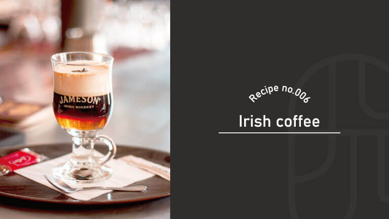 A layered Irish coffee in a footed glass mug branded Jameson, with cream floating on top, served on a plate with a spoon