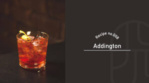 A glass of the Addington cocktail on ice, garnished with a green olive, served in a dimly lit bar setting