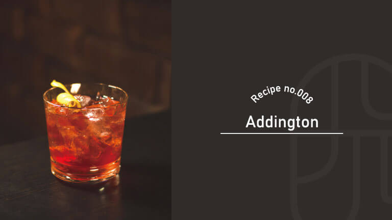 A glass of the Addington cocktail on ice, garnished with a green olive, served in a dimly lit bar setting