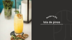 A tall glass of orange-colored Isla de Pinos cocktail garnished with rosemary and dried citrus slices, served on a coaster