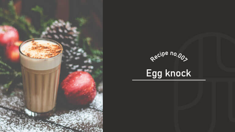 A glass of creamy eggnog cocktail topped with foam and nutmeg, surrounded by pinecones and red apples
