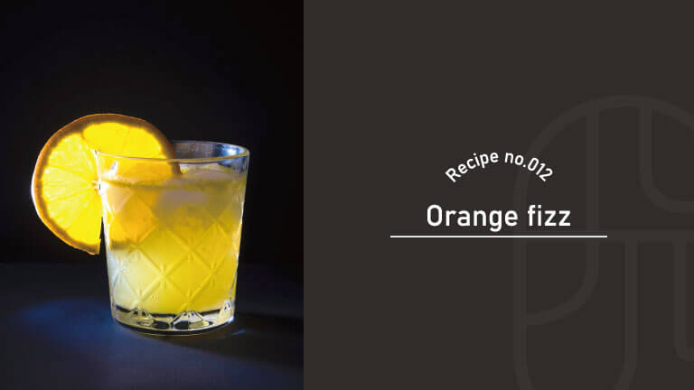 A glass of orange fizz cocktail garnished with an orange wheel, lit dramatically against a dark background