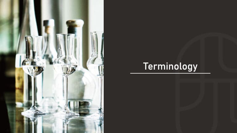Clear glasses of spirits beside bottles and a decanter, reflected on a polished bar counter with soft window light