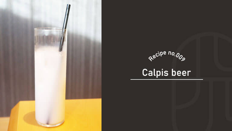 A tall glass of milky-white Calpis beer cocktail with a straw, served on a yellow table in soft light