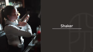 A bartender in glasses and suspenders holding a cocktail shaker up near her shoulder at a bar counter, with "Shaker" text overlay