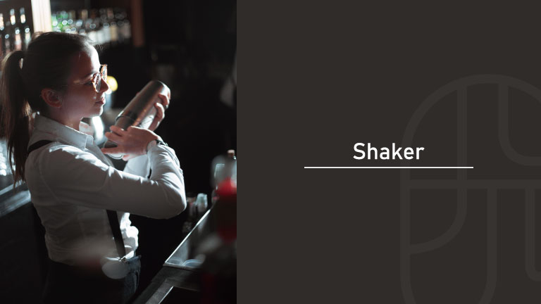 A bartender in glasses and suspenders holding a cocktail shaker up near her shoulder at a bar counter, with "Shaker" text overlay