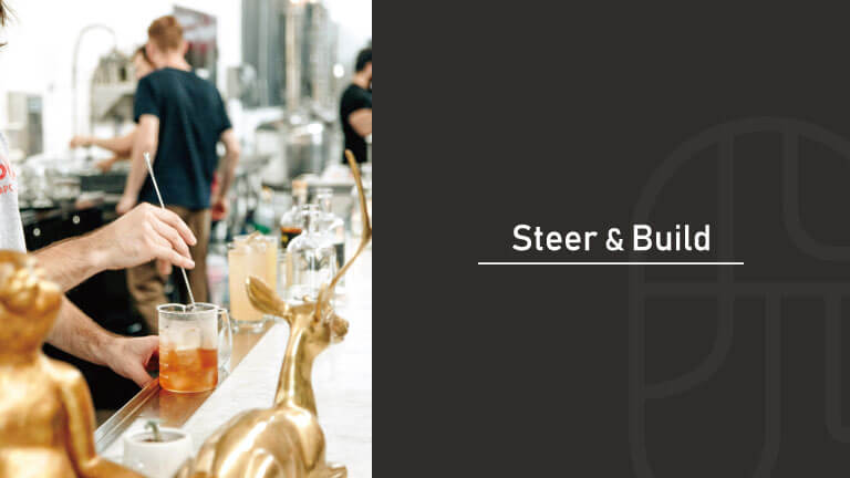 A bartender stirring a cocktail with a bar spoon at a bar counter decorated with golden animal figurines, with "Steer & Build" text overlay