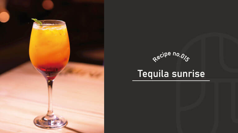 A tequila sunrise cocktail with its signature red-to-orange gradient, garnished with mint, served in a wine glass on a wooden table