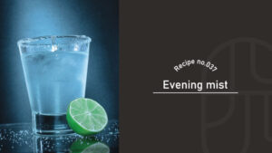 A salt-rimmed glass of clear Evening Mist cocktail with a lime half beside it, lit against a moody blue background