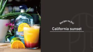 A layered orange and red California Sunset cocktail garnished with orange, beside a tequila bottle and lime