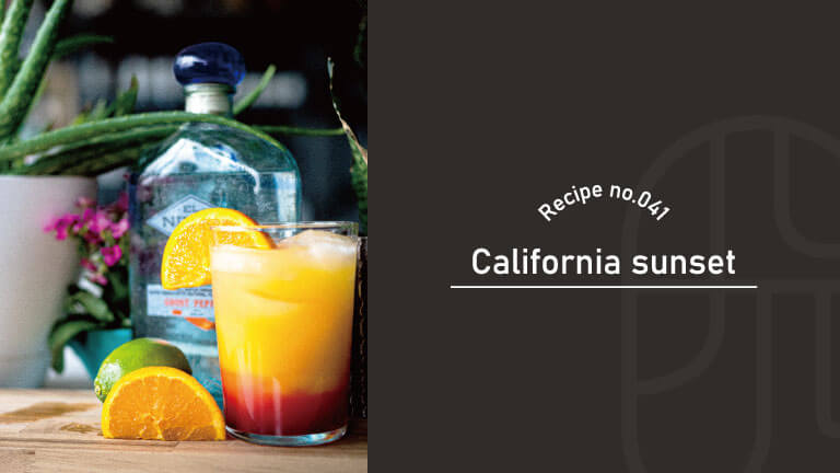 A layered orange and red California Sunset cocktail garnished with orange, beside a tequila bottle and lime