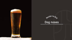 A tall glass of amber Dog Noses cocktail with a foamy white head, set against a dark background