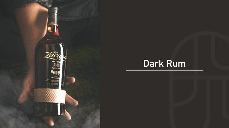 A hand holding a bottle of Ron Zacapa 23 dark rum against a dark, smoky background