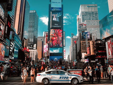 A busy street scene in Times Square, New York City, with illuminated billboards, an NYPD car, and pedestrians crossing