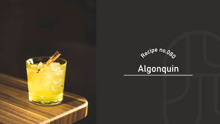 A pale yellow Algonquin cocktail served over ice in a rocks glass, garnished with a cinnamon stick, on a wooden bar counter