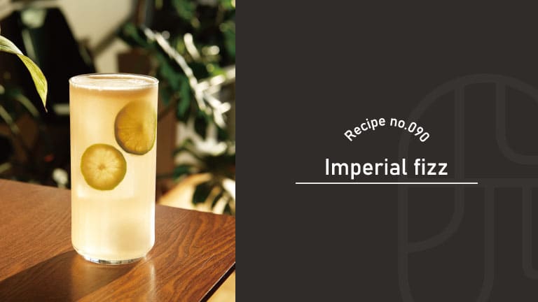 A pale golden Imperial Fizz cocktail served in a tall glass with lime slices, set on a wooden table with greenery in the background