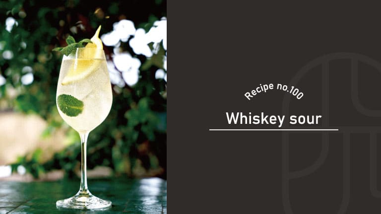 A stemmed glass of pale golden Whiskey Sour cocktail garnished with a lemon wheel and mint leaves, set outdoors against greenery and white flowers, beside a "Recipe no.100 Whiskey sour" title card