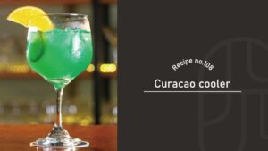 A vivid green Curacao Cooler in a stemmed goblet garnished with an orange wheel and cucumber, on a wooden bar counter, beside a "Recipe no.108 Curacao cooler" title card