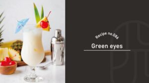 A creamy white Green Eyes cocktail in a hurricane glass, garnished with pineapple wedges, a maraschino cherry, and a blue cocktail umbrella