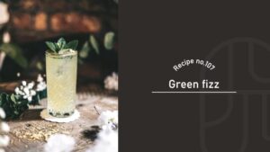 A pale green Green Fizz cocktail garnished with mint in a cut-crystal glass, on a wooden table with white flowers, beside a "Recipe no.107 Green fizz" title card