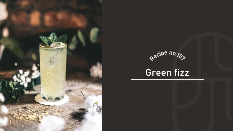 A pale green Green Fizz cocktail garnished with mint in a cut-crystal glass, on a wooden table with white flowers, beside a "Recipe no.107 Green fizz" title card